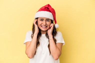Genç beyaz kadın Noel 'i kutluyor. Sarı arka planda izole edilmiş. Kulaklarını elleriyle kapatıyor..