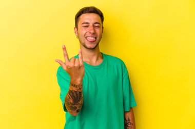 Sarı arka planda izole edilmiş dövmeleri olan genç beyaz bir adam parmaklarıyla taş hareketi gösteriyor.