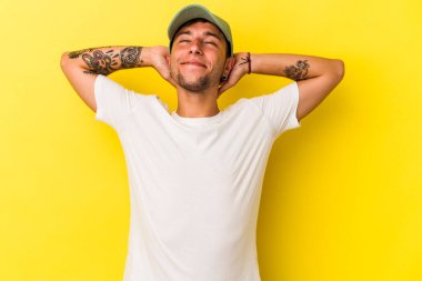 Sarı arka planda izole edilmiş, elleri başının arkasında kendinden emin dövmeleri olan genç, beyaz bir adam..