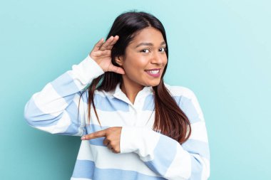 Genç Venezuelalı bir kadın mavi arka planda izole edilmiş bir şekilde bir dedikoduyu dinlemeye çalışıyor..