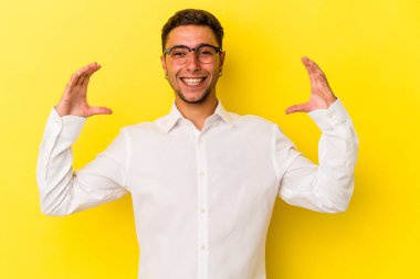 Sarı arka planda izole edilmiş beyaz dövmeli genç bir adam avuçlarıyla bir şey tutuyor ve kameraya sunuyor..