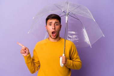 Mor arka planda izole edilmiş şemsiyeli genç beyaz adam yan tarafı işaret ediyor.