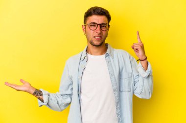 Sarı arka planda izole edilmiş dövmeleri olan genç beyaz bir adam elinde bir ürün taşıyor..