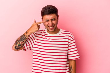 Sarı arka planda elleri kulaklarında, dövmeleri olan genç, beyaz bir adam..