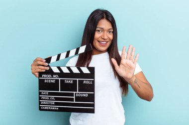 Genç Venezuelalı bir kadın mavi arka planda izole edilmiş bir klempon tutuyor. Beş numarayı parmaklarıyla gülümsüyor..