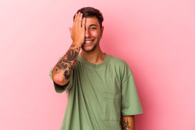 Sarı arka planda dövmeleri olan beyaz bir adam yüzünün yarısını avuç içiyle kaplayarak eğleniyor..