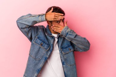 Sarı arka planda izole edilmiş dövmeleri olan beyaz genç bir adam parmaklarıyla kameraya göz kırpıyor ve yüzünü kapatmaktan utanıyor..