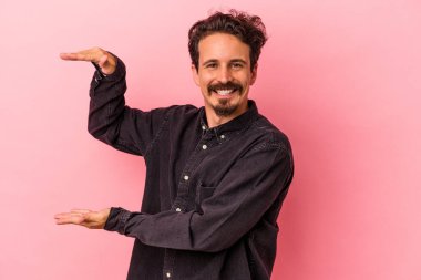 Pembe arka planda izole edilmiş genç beyaz adam işaret parmaklarıyla küçük bir şey tutuyor, gülümsüyor ve kendinden emin..