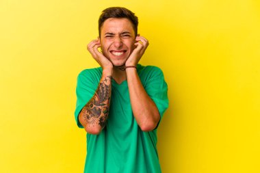 Sarı arka planda elleri kulaklarında, dövmeleri olan genç, beyaz bir adam..