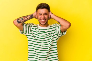 Sarı arka planda elleri kulaklarında, dövmeleri olan genç, beyaz bir adam..