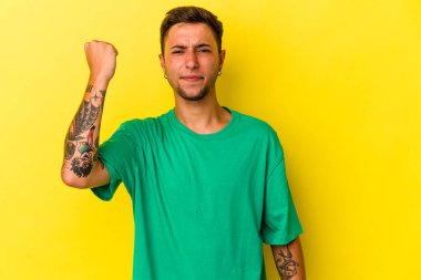 Sarı arka planda izole edilmiş dövmeleri olan genç beyaz bir adam kameraya yumruk gösteriyor, agresif bir yüz ifadesi var..