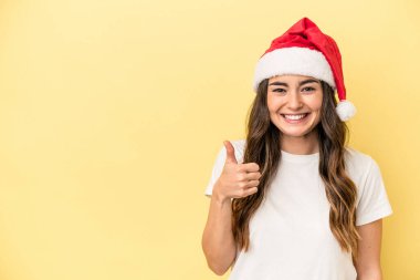 Genç beyaz kadın Noel 'i kutluyor sarı arka planda izole edilmiş gülümsüyor ve başparmağını kaldırıyor.