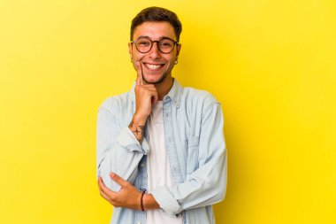 Sarı arka planda izole edilmiş, mutlu ve kendinden emin gülümseyen, elleriyle çenesine dokunan beyaz dövmeli genç bir adam..