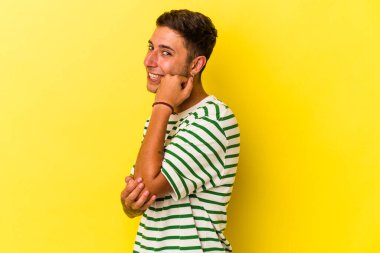 Sarı arka planda izole edilmiş, mutlu ve kendinden emin gülümseyen, elleriyle çenesine dokunan beyaz dövmeli genç bir adam..