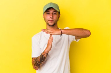 Sarı arka planda izole edilmiş dövmeleri olan genç beyaz bir adam, bir zaman aşımı hareketi gösteriyor..