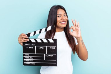 Genç Venezuelalı bir kadın mavi arka planda izole edilmiş bir el çırpma tahtası tutuyor. Neşeli ve kendinden emin..