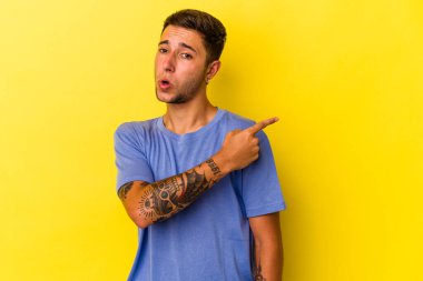 Sarı arka planda izole edilmiş, gülümseyen ve işaret eden, boş alanda bir şeyler gösteren beyaz dövmeli genç bir adam..