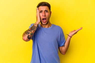 Sarı arka planda izole edilmiş dövmeleri olan beyaz genç bir adam, avuç içi fotokopi alanını elinde tutmaktan etkilenmiş..