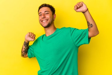 Sarı arka planda, özel bir günü kutlayan dövmeli, genç, beyaz bir adam atlıyor ve enerjiyle kollarını kaldırıyor..