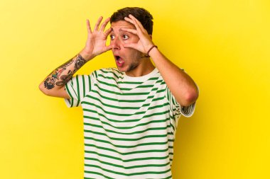 Sarı arka planda izole edilmiş dövmeleri olan beyaz genç bir adam başarılı bir fırsat yakalamak için gözlerini açık tutuyor..