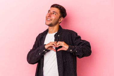 Sarı arka planda izole edilmiş dövmeleri olan genç beyaz bir adam gülümsüyor ve elleriyle kalp şeklini gösteriyor..