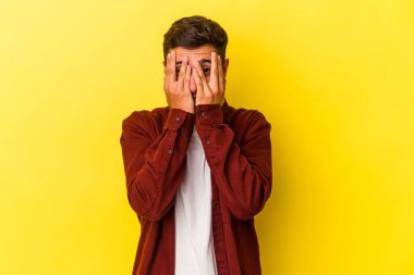 Sarı arka planda izole edilmiş dövmeleri olan beyaz genç bir adam parmaklarıyla kameraya göz kırpıyor ve yüzünü kapatmaktan utanıyor..