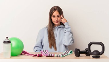 Beyaz arka planda izole edilmiş spor ekipmanlarıyla masada oturan beyaz tenli genç bir kadın şakağı parmağıyla işaret ediyor, düşünüyor ve bir göreve odaklanıyor..