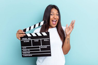 Genç Venezuelalı bir kadın mavi arka planda izole edilmiş bir el çırpma tahtası tutuyor. Hoş bir sürpriz alıyor, heyecanlı ve el kaldırıyor..