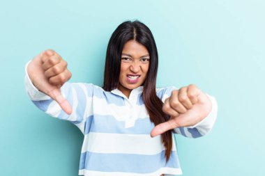 Genç Venezuelalı bir kadın mavi arka planda izole edilmiş, baş parmağını aşağı gösteren ve hoşlanmadığını ifade eden.
