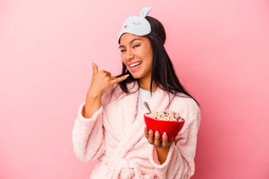 Pijama giyen genç Latin bir kadın pembe arka planda izole edilmiş bir kase mısır gevreği tutarken parmaklarıyla bir cep telefonu hareketi gösteriyor..