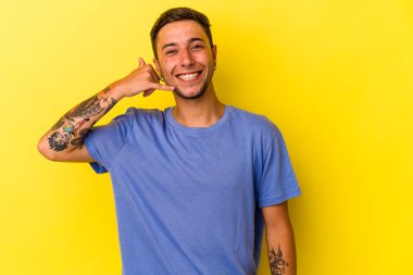 Sarı arka planda izole edilmiş dövmeleri olan genç beyaz bir adam parmakları olan bir cep telefonu hareketi gösteriyor..