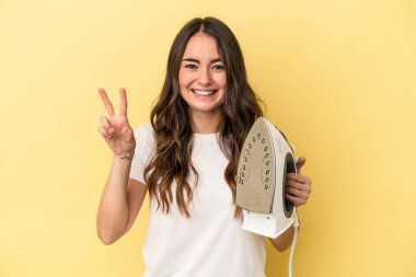 Young caucasian woman holding an iron isolated on yellow background showing number two with fingers.