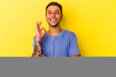 Sarı arka planda izole edilmiş dövmeleri olan beyaz bir genç göz kırpıyor ve elleriyle iyi bir jest yapıyor..