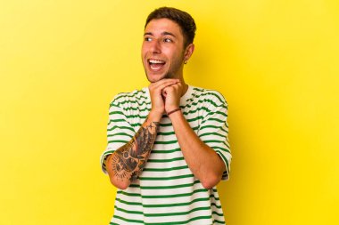 Sarı arka planda dövmeleri olan genç beyaz adam ellerini çenesinin altında tutuyor ve mutlu bir şekilde yan tarafa bakıyor..