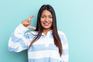 Genç Venezuelalı bir kadın mavi arka planda izole edilmiş. Elinde parmakları olan, gülümseyen ve kendinden emin bir şey var..