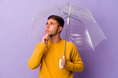 Mor arka planda şemsiyesi olan beyaz bir adam şüpheli ve şüpheci bir ifadeyle yana bakıyor..