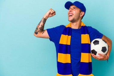 Genç futbolcu elinde mavi arka planda izole edilmiş bir top tutuyor. Zaferden sonra yumruğunu kaldırıyor, kazanan konsept..