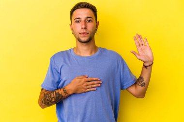 Sarı arka planda izole edilmiş dövmeleri olan beyaz genç bir adam yemin ediyor, elini göğsüne koyuyor..