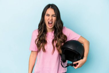 Young caucasian woman holding a motor bike helmet isolated on blue background screaming very angry and aggressive.