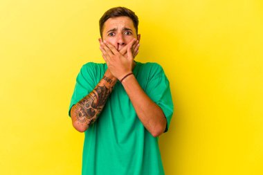 Sarı arka planda dövmeleri olan beyaz bir genç adam elleri ile ağzını kapatırken şoka uğramış..