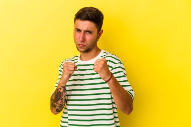 Sarı arka planda izole edilmiş dövmeleri olan genç beyaz bir adam kameraya yumruk gösteriyor, agresif bir yüz ifadesi var..