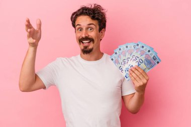 Pembe arka planda banknotları elinde tutan beyaz bir genç hoş bir sürpriz alıyor, heyecanlı ve el kaldırıyor..