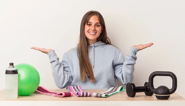 Young caucasian woman sitting at a table with sport equipment isolated on white background makes scale with arms, feels happy and confident.