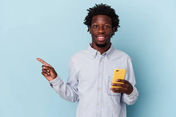 年轻的非洲裔美国人拿着被蓝色背景隔离的手机 微笑着指着旁边 在空白的空间里展示着一些东西 — 图库照片
