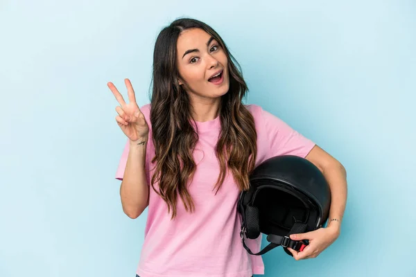 Young caucasian woman holding a motor bike helmet isolated on blue background joyful and carefree showing a peace symbol with fingers.