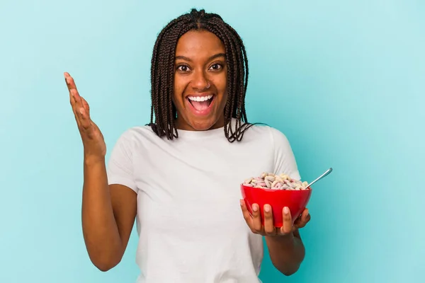 年轻的非洲裔美国女人手里拿着一碗蓝色背景的麦片 惊喜极了 兴奋地举起双手 — 图库照片