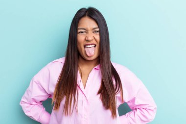 Genç Venezuelalı bir kadın mavi arka planda izole edilmiş komik ve dost canlısı bir dil kullanıyor..