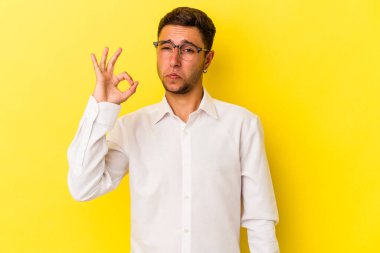 Sarı arka planda izole edilmiş dövmeleri olan beyaz bir genç göz kırpıyor ve elleriyle iyi bir jest yapıyor..