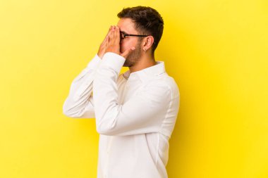 Sarı arka planda izole edilmiş dövmeleri olan genç beyaz bir adam elleriyle gözlerini kapatmaktan korkuyor..