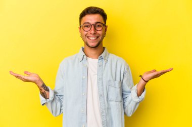 Sarı arka planda dövmeleri olan genç beyaz adam kollarıyla ölçek çiziyor, mutlu ve kendinden emin hissediyor..